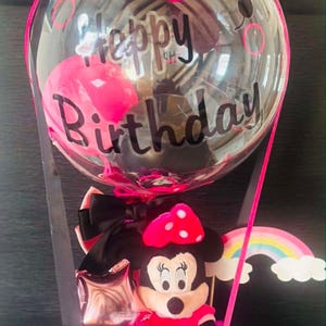 May include: A clear balloon with the text Happy Birthday in black script, topped with a pink bow. Below, a plush mouse character with a pink polka-dot bow sits in a white cylindrical box decorated with black dots, accented by a rainbow cutout.
