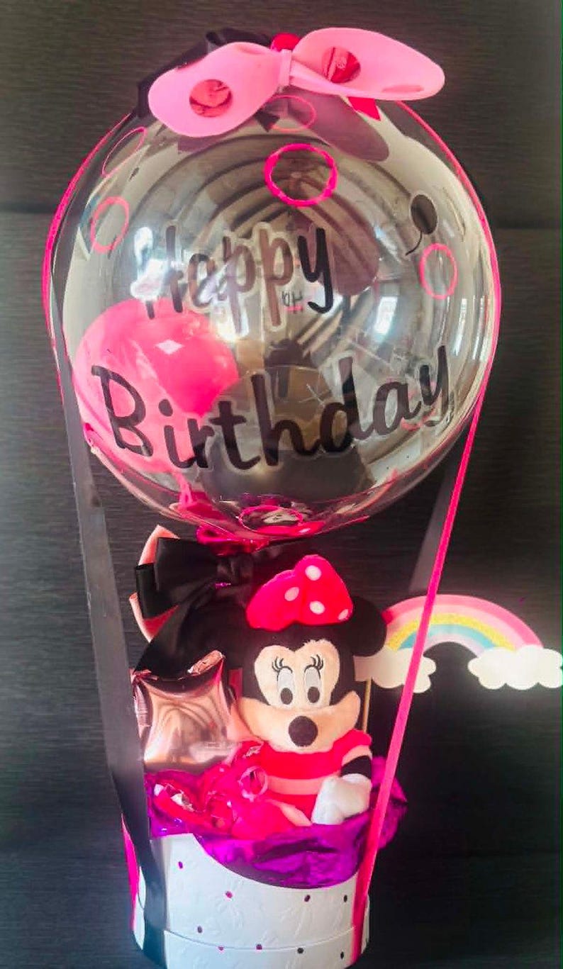 May include: A clear balloon with the text Happy Birthday in black script, topped with a pink bow. Below, a plush mouse character with a pink polka-dot bow sits in a white cylindrical box decorated with black dots, accented by a rainbow cutout.