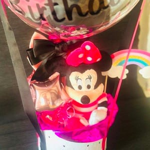 May include: A gift arrangement featuring a plush Minnie Mouse character with a pink polka dot bow, nestled in a white round box with black accents. A clear balloon with the word Birthday printed in black script sits above, accented by a rainbow cutout.