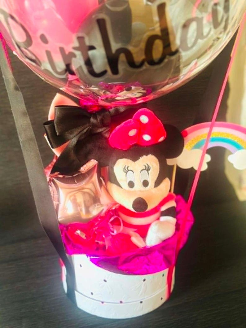 May include: A gift arrangement featuring a plush Minnie Mouse character with a pink polka dot bow, nestled in a white round box with black accents. A clear balloon with the word Birthday printed in black script sits above, accented by a rainbow cutout.