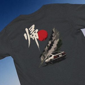 Könnte beinhalten: Ein dunkelgraues T-Shirt mit einem Grafikdruck auf der Rückseite. Das Design zeigt ein weißes japanisches Schriftzeichen, einen roten Kreis und einen weißen Sportwagen von hinten vor einem Hintergrund aus grauen Pinselstrichen.
