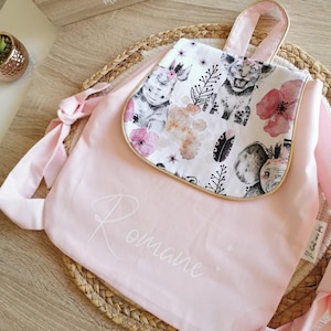 Peut inclure: Un sac à dos rose avec un rabat orné d'un motif aquarelle d'animaux et de fleurs. Le sac à dos a une poignée et des bretelles. Le nom "Romane" est écrit en cursive sur le devant. Les mots "Beautiful Moments" sont sur une boîte en bois.