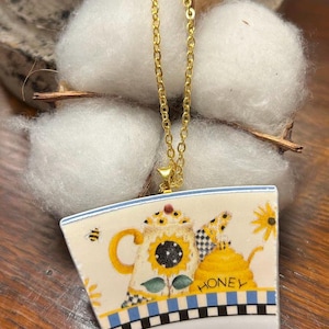 May include: A gold-colored chain necklace with a pendant featuring a teapot design. The pendant has a sunflower and honey pot illustration with the word "HONEY". The background includes cotton and a wooden surface.