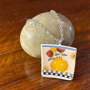 May include: A square pendant necklace with a floral design. The pendant features a yellow vase with red and yellow flowers. The border has a blue and white checkered pattern. The necklace chain is silver.