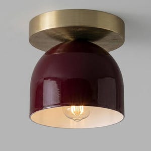 May include: A modern ceiling light fixture with a burgundy dome-shaped shade and a brass-colored circular base. The light has a visible bulb and a white interior. The fixture is designed for indoor use.