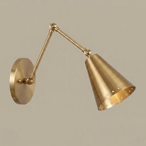 Op de afbeelding: Een messingkleurige wandlamp met een verstelbare arm en een conische kap. De lamp heeft een ronde basis en kleine gaatjes rond de onderkant van de kap. Het ontwerp doet denken aan vintage industriële verlichting.