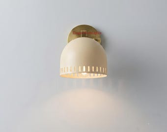 May include: A wall sconce featuring a dome-shaped, cream-colored metal shade with a series of vertical rectangular cutouts along the bottom edge. The fixture is mounted to the wall with a circular, brushed brass-toned metal backplate.