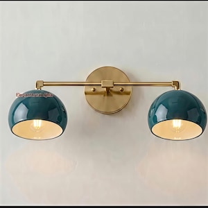 Può includere: Una lampada da parete con una barra orizzontale in ottone fissata a una piastra circolare in ottone. Due paralumi a cupola color verde acqua sono posizionati a ciascuna estremità, ognuno contenente una lampadina. Il testo "ElepantAuraLights" è visibile.