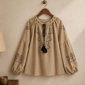 May include: A beige long-sleeve blouse featuring black floral embroidery around the neckline and on the sleeves. The garment has a round neckline with a black drawstring tie finished with two black tassels hanging at the centre of the chest.