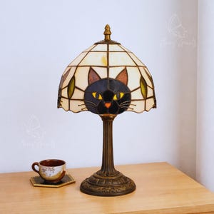 Könnte beinhalten: Tischlampe aus Buntglas mit einem schwarzen Katzenmotiv auf dem Lampenschirm. Die Lampe hat einen Sockel und einen Stiel in Bronzefarbe. Eine kleine Teetasse und Untertasse stehen auf einer Holzoberfläche.