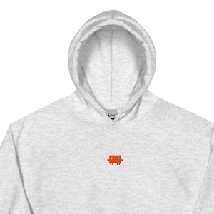 May include: A light heather gray hooded sweatshirt featuring a small, orange, pixelated square character embroidered on the center of the chest. The garment includes a drawstring hood, long sleeves with ribbed cuffs, and a large front kangaroo pocket.