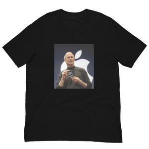 Può includere: Una t-shirt nera a maniche corte con una stampa grafica centrale di Steve Jobs che indossa una maglia a collo alto nera, mentre tiene in mano un piccolo smartphone davanti a un grande logo Apple bianco su uno sfondo grigio scuro.