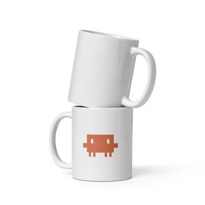 May include: Two white ceramic coffee mugs stacked on top of each other. The bottom mug features a pixelated orange graphic. Both mugs have a curved handle and a smooth, glossy finish. The mugs are set against a white background.