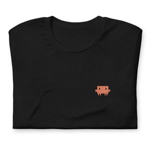 May include: Black t-shirt featuring a small, pixelated, orange-brown couch emblem on the chest. The shirt is folded, displaying the neckline and the soft material. A simple, minimalist design.