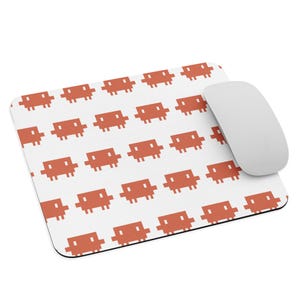 May include: A rectangular mouse pad with a white background and a repeating pattern of orange pixelated alien figures. A smooth, light gray computer mouse rests on the right side of the mouse pad, partially covering the pattern of the pixelated figures.