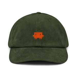 May include: An olive green corduroy baseball cap featuring a small, embroidered orange pixelated alien graphic centered on the front panel. The cap has a structured crown, a curved brim, and a classic six-panel design with matching top stitching.