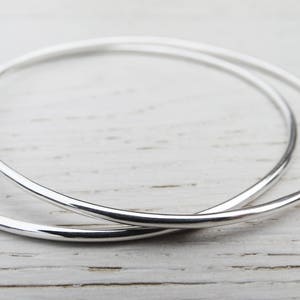 May include: Two silver hoop earrings, one slightly larger than the other, are shown on a white wooden surface.