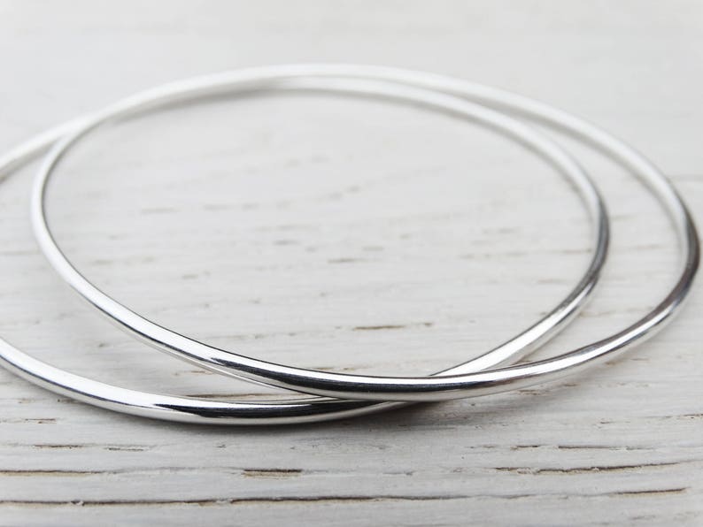 May include: Two silver hoop earrings, one slightly larger than the other, are shown on a white wooden surface.