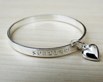 May include: A silver bangle bracelet with the name "Scarlett" engraved on it. A small silver heart charm hangs from the bangle.