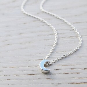 May include: A silver crescent moon pendant necklace on a delicate silver chain. The necklace is displayed on a light wood surface.