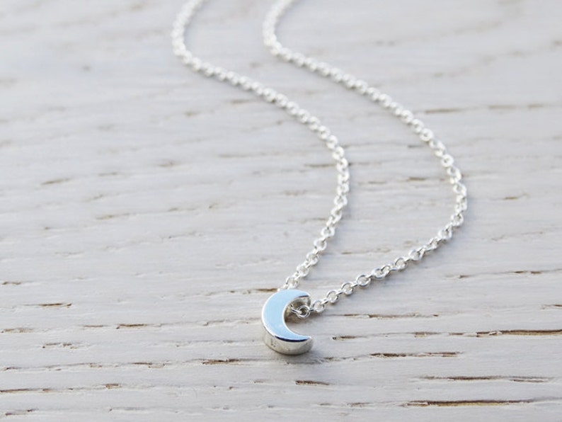May include: A silver crescent moon pendant necklace on a delicate silver chain. The necklace is displayed on a light wood surface.