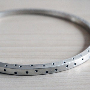 May include: A silver bangle bracelet with a series of evenly spaced, small, dark holes around the outer edge. The bracelet has a matte finish and a simple, modern design. The jewelry is displayed on a light-coloured wooden surface.