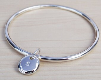 May include: A silver bangle bracelet with a round silver charm that has the letter "S" engraved on it.
