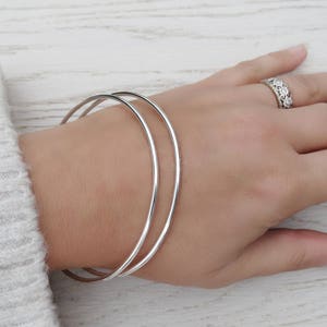 May include: Two silver bangle bracelets stacked on a person's wrist. The bracelets are simple and smooth.