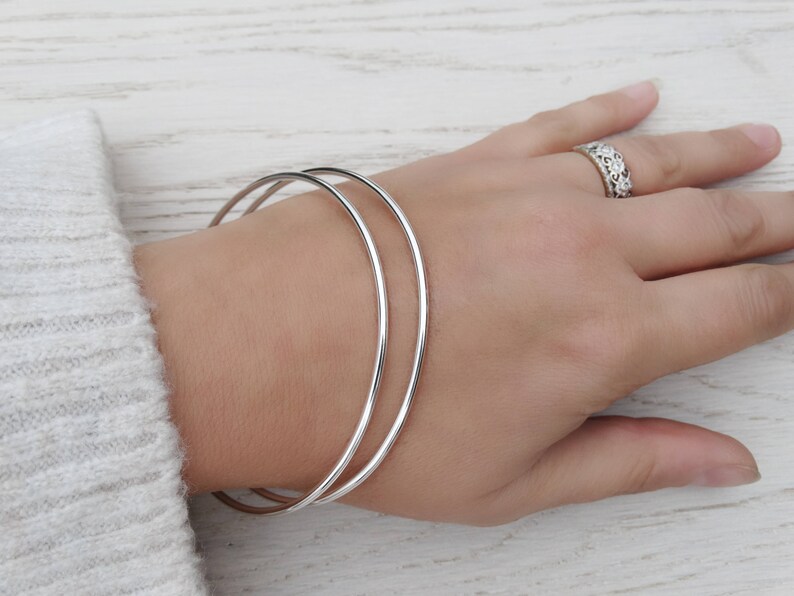 May include: Two silver bangle bracelets stacked on a person's wrist. The bracelets are simple and smooth.