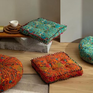 May include: Square floor cushions featuring intricate patterns in vibrant teal and orange tones. Each cushion is finished with small pom-pom trim along the edges and includes tufted detailing on the surface for a textured appearance.