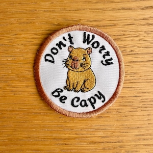 Könnte beinhalten: Ein runder, bestickter Aufnäher mit weißem Hintergrund und braunem Rand. In der Mitte befindet sich eine Illustration eines beigen Wasserschweins, umgeben von dem schwarzen Sticktext "Don't Worry Be Capy" in kreisförmiger Anordnung.