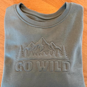 May include: A dusty blue crewneck sweatshirt featuring an embroidered mountain range and forest silhouette above the text GO WILD. The design is stitched in the same dusty blue thread as the fabric, creating a subtle, raised, tonal texture.