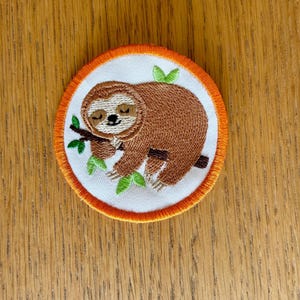 Könnte beinhalten: Gestickter Aufnäher mit einem braunen Faultier, das auf einem Ast mit grünen Blättern ruht. Das Faultier befindet sich auf einem weißen Kreis mit orangefarbenem Rand. Ideal für Kleidung oder Accessoires.