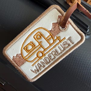 Könnte beinhalten: Besticktes Gepäckanhänger mit cremefarbenem Hintergrund, mit einem braunen und goldenen Wohnwagen-Design und dem Wort "WANDERLUST". Der Anhänger hat einen braunen Lederriemen.