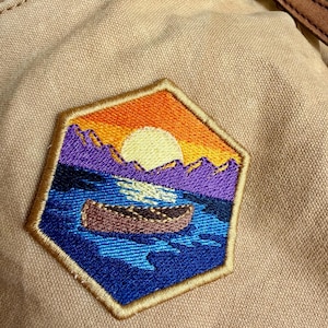Könnte beinhalten: Sechseckiger, gestickter Aufnäher mit einem Sonnenuntergang über einem See mit einem Kanu. Der Aufnäher hat einen goldenen Rand. Der Himmel ist orange und gelb, die Berge sind lila und das Wasser ist blau. Der Aufnäher ist auf einem beigen Stoff angebracht.