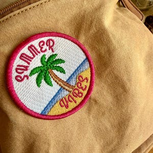 Könnte beinhalten: Ein beigefarbener Canvas-Rucksack mit einem kreisförmigen, gestickten Aufnäher. Der Aufnäher zeigt die Worte "Summer Vibes" in Rosa, umgeben von einer Palme an einem Strand mit blauem Wasser und gelbem Sand.
