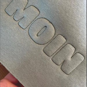 Könnte beinhalten: Ein salbeigrünes Kleidungsstück aus Stoff mit dem Wort MOIN in erhabener, gepolsterter Optik. Die Buchstaben sind mit dunklen Nähten umrandet, was einen dreidimensionalen Effekt auf der glatten, matten Oberfläche des Textilmaterials erzeugt.