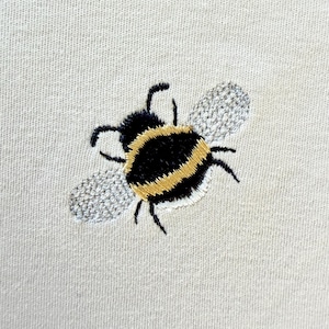 Könnte beinhalten: Gestickte Hummel in Schwarz, Gelb und Weiß auf hellbeigem Stoff. Die Biene hat detaillierte Flügel und einen strukturierten Körper, was ein charmantes Insektenmotiv ergibt. Die Stickerei befindet sich wahrscheinlich auf einem Hemd oder Textil.