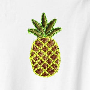 Puede incluir: Diseño de piña bordada en tonos marrón, verde y amarillo. La piña tiene un aspecto texturizado con un patrón de punto de cruz. Las hojas son de color verde oscuro y el cuerpo es de un verde amarillento más claro.