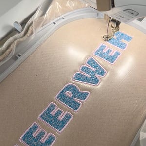 Könnte beinhalten: Eine weiße Stickmaschine stickt das Wort MEERWER mit blauem, strukturiertem Garn und einer hellrosa Umrandung auf einen beigen Stoff, der in einem weißen Kunststoff-Stickrahmen eingespannt ist. Die Nadel befindet sich am Ende des Buchstabens H.