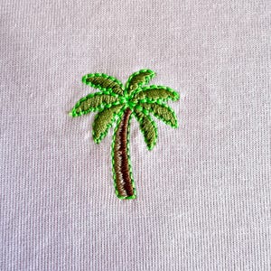 Könnte beinhalten: Nahaufnahme eines hellrosa Stoffes mit einem gestickten Palmenmotiv. Die Palme hat einen braunen Stamm und grüne Wedel, die mit leuchtend grünen Fäden umrandet sind. Die Stickerei ist detailliert und strukturiert den Stoff.