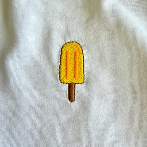 Op de afbeelding: Een wit t-shirt met een geborduurd geel ijslolly-ontwerp. De ijslolly heeft oranje details en een bruin stokje. Het ontwerp is gecentreerd op het shirt en biedt een eenvoudige, zomerse uitstraling.