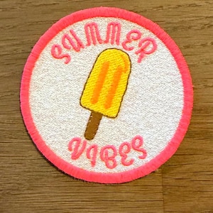 Könnte beinhalten: Runder Aufnäher mit weißem Zentrum, leuchtend rosa Rand und den Worten "SUMMER VIBES" in rosa Schrift. Ein gelbes Eis am Stiel mit orangefarbenen Streifen und einem braunen Stiel befindet sich in der Mitte.