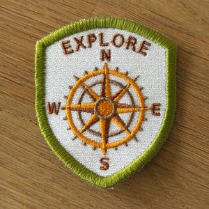 Könnte beinhalten: Ein schildförmiger, bestickter Aufnäher mit weißem Hintergrund und lindgrünem Rand. Der Aufnäher zeigt in der Mitte eine braune Kompassrose, darüber das Wort EXPLORE in einem Bogen sowie die Himmelsrichtungen N, S, E und W.