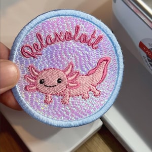 Könnte beinhalten: Ein runder, bestickter Aufnäher mit einem rosa Axolotl-Motiv auf einem schimmernden, irisierenden violetten Hintergrund. Der Schriftzug Relaxalotl ist in rosa Schreibschrift über dem Axolotl gestickt, umrandet von einem hellblauen Rand.
