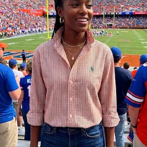 May include: A long-sleeved button-down shirt featuring a vertical striped pattern in shades of orange, white, and blue. The shirt has a ruffled collar, a buttoned front, and a small green embroidered logo on the left chest area, paired with blue denim jeans.