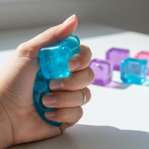 Puede incluir: Una mano aprieta una bola antiestrés translúcida azul en forma de cubo. En el fondo se ven varios cubos de colores, incluidos morados y rosas. La bola antiestrés está hecha de un material similar al gel.