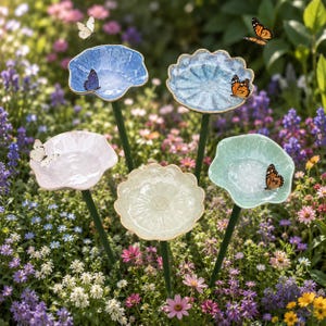 May include: Five decorative flower-shaped bird baths in a garden. The bird baths are in shades of blue, pink, green, and cream, with gold edging. Butterflies are resting on the bird baths. The bird baths are mounted on green stems.