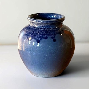 May include: A small, rounded ceramic vase featuring a glossy glaze that transitions from a deep, dark blue at the rim to a lighter, mottled blue and earthy brown tone toward the base. The vessel has a short, narrow neck and a defined, rounded lip.