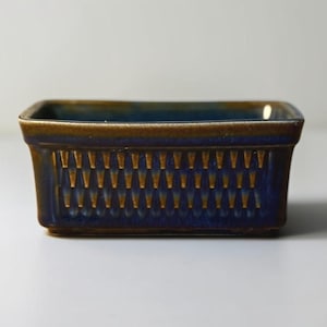 May include: A rectangular ceramic dish featuring a deep blue glaze with brown accents along the rim and base. The exterior walls are decorated with a repeating vertical pattern of small, recessed triangular indentations in a golden brown hue.
